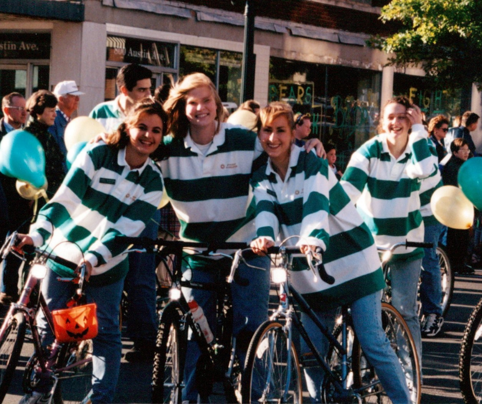 4 college students in green and white long sleeve shirts on top of bikes for Baylor homecoming. The girl on the far left is holding an orange jack-o-lantern bucket.