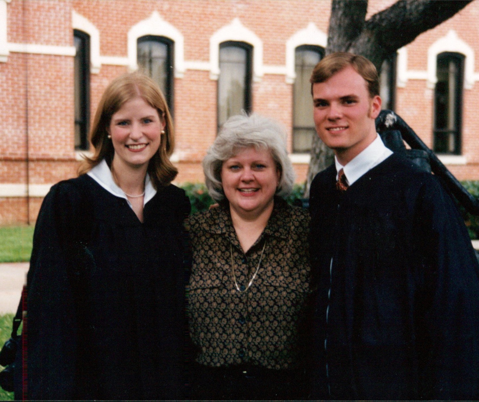 A young woman, older woman, and man are standing outside of a brick building in what look like graduation robes