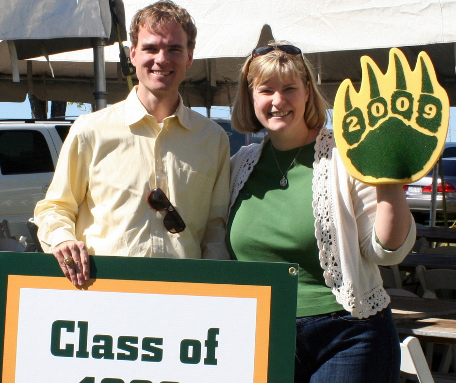 A man and a woman stand by a class sign, the woman is wearing a foam bear claw with 2009 on it