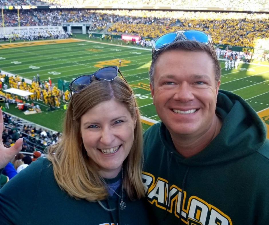 A man and a woman are at a football game, they are smiling and wearing sunglasses on top of their heads and the man is wearing a green Baylor sweatshirt